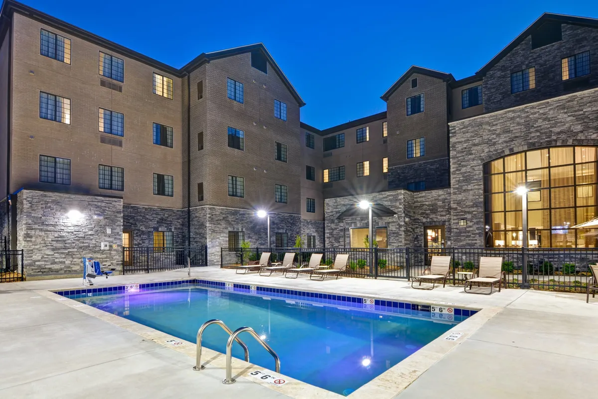 This image features an inviting outdoor swimming pool surrounded by lounge chairs and the building's facade, with beautifully landscaped grounds and a glowing sunset in the background. Several guests can be seen relaxing by the pool and socializing, evoking a relaxed and fun atmosphere.