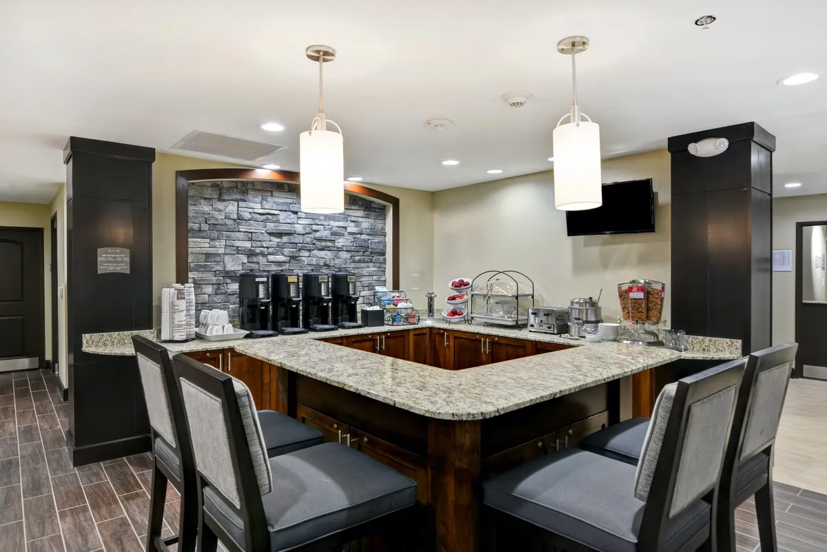 A welcoming interior scene featuring a granite countertop with a coffee station, adjacent to a decorative stone wall and cozy seating area. The space feels open, bright, and designed for guests to gather for breakfast or quick snacks.