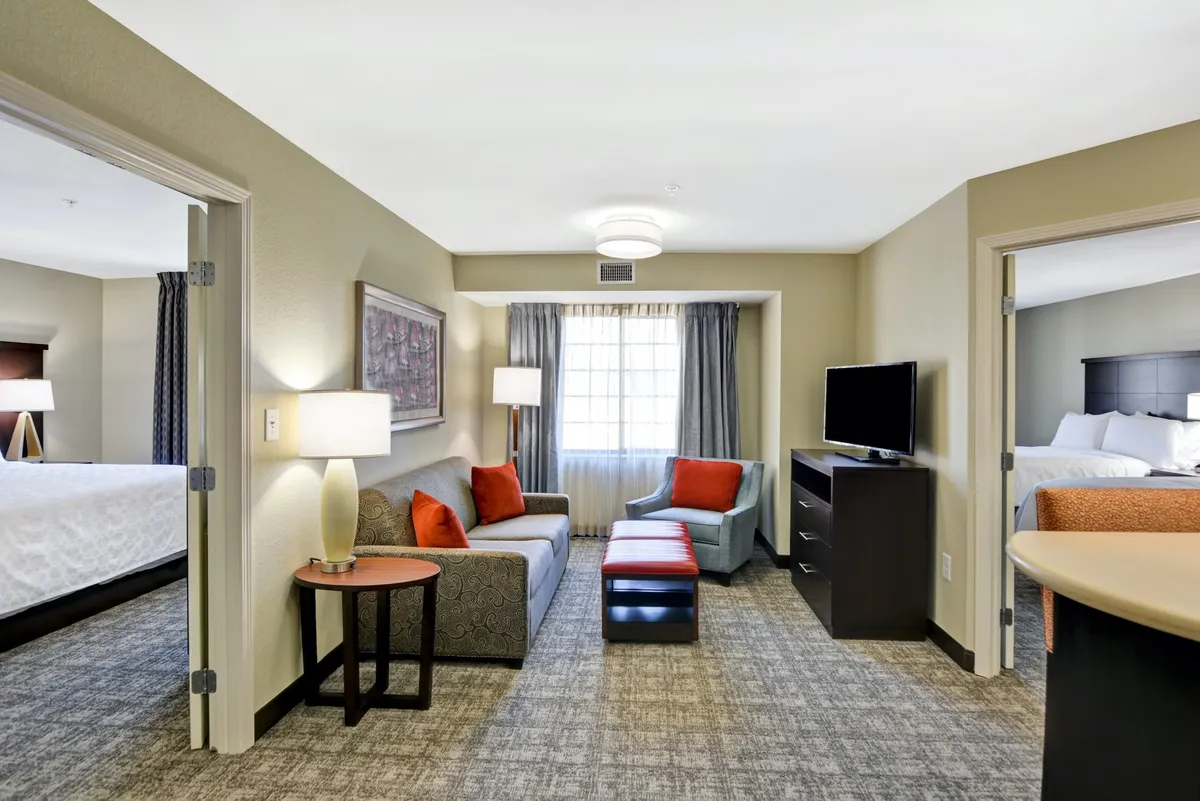 A cozy living room featuring a plush sofa with vibrant orange cushions, a stylish coffee table, a warm lamp, a flat-screen TV, and a sunlit window with elegant curtains. The setting invites guests to relax, socialize, or enjoy a quiet moment by the window.