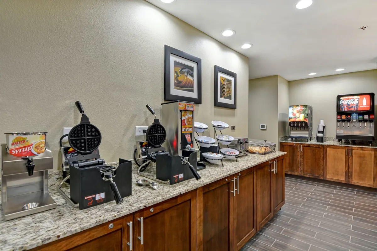A friendly and relaxed breakfast station featuring waffle makers, a syrup dispenser, beverage station, and snack bowls, surrounded by colorful, inviting decor. The setup is designed to encourage guests to create their own breakfasts and engage with others.