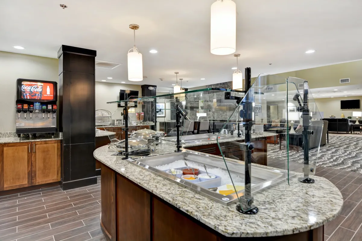 A bright self-service breakfast area showcasing a granite countertop lined with a food display and beverage station, illuminated by modern lighting fixtures. The setup suggests a casual, welcoming environment where guests can enjoy breakfast and interact.