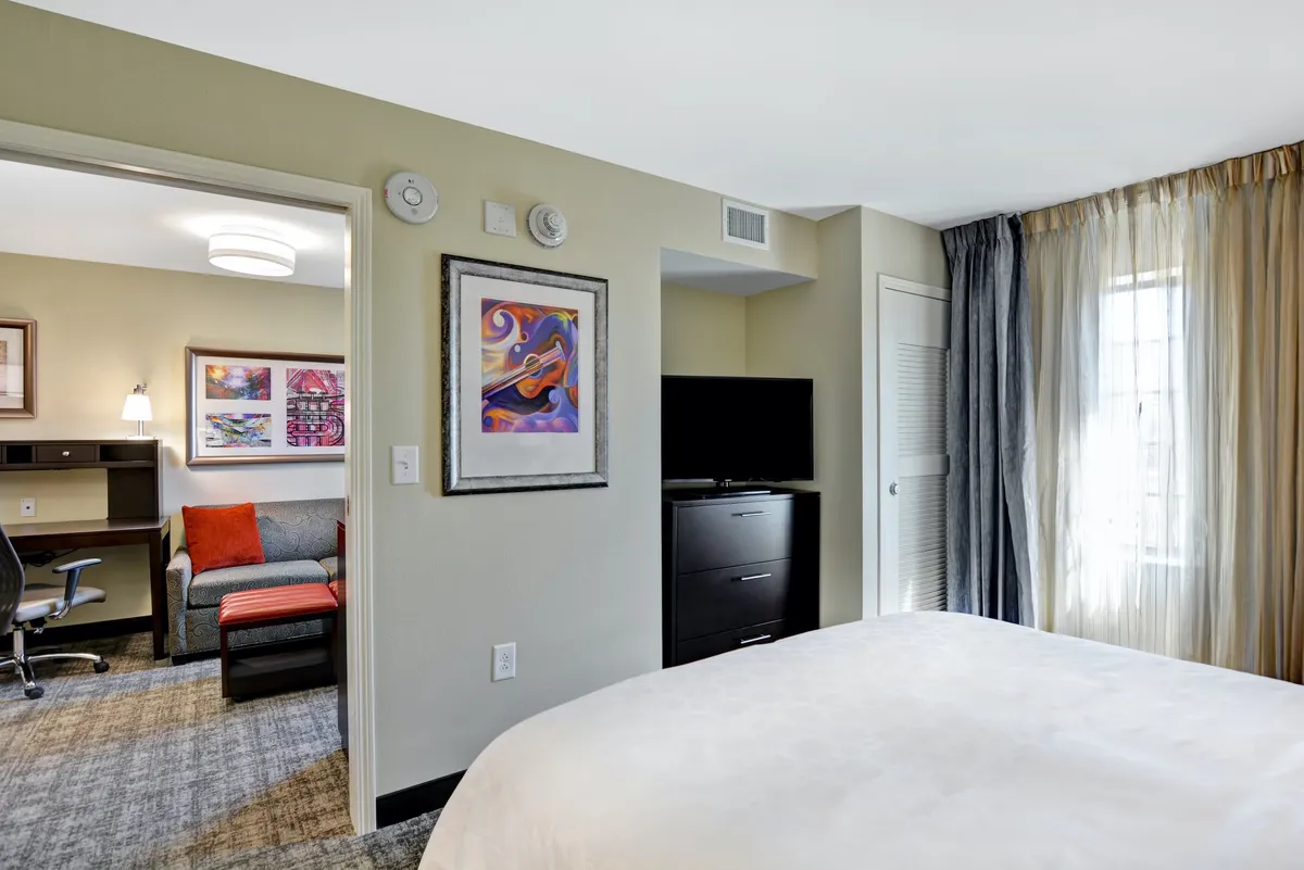 This image features a serene bedroom with a plush bed, elegant artwork above the headboard, a comfortable sofa in the corner, and a functional desk, all arranged to provide a cozy and versatile environment. The clean lines and soft lighting emphasize restfulness and comfort, while the decor conveys a sense of personal retreat.