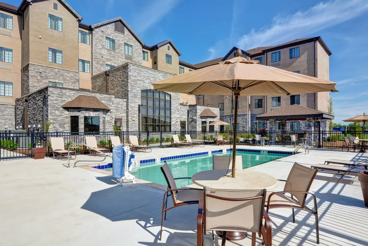 A bright, cheerful view of the outdoor swimming pool with ample seating, umbrellas, and the hotel building in the background. The area is set up for sunbathing, swimming, and casual outdoor dining, making it attractive for families to gather and enjoy leisure time together.