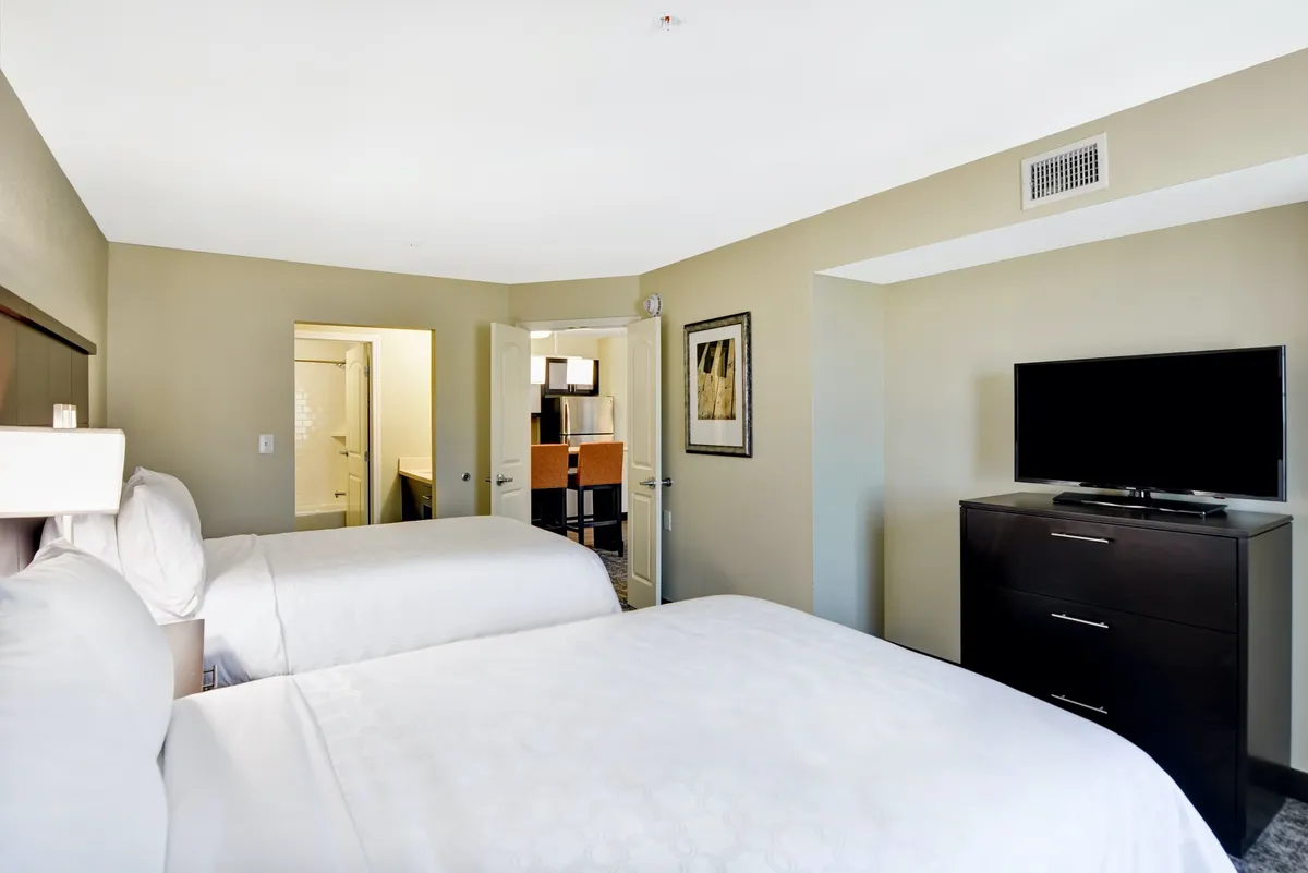 A spacious, bright bedroom setup featuring two comfortable beds, inviting linens, a TV for entertainment, and tasteful wall art. The room's layout offers easy access to adjoining areas such as the kitchen and bathroom, making it ideal for families, friends, or colleagues sharing the space.